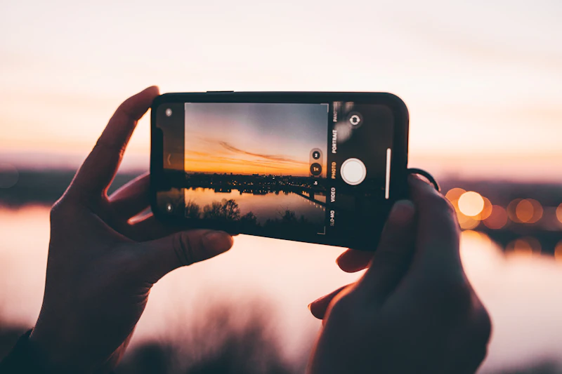 Two hands holding a smartphone up to photograph a golden sunset over water