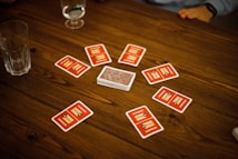 A wooden table with several playing cards arranged in a circle around a stack of cards in the center. Each card has a red back with text and illustrations. Two drinking glasses, one with a clear liquid, are also on the table, with a person's arm visible in the background.