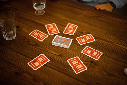 A wooden table with several playing cards arranged in a circle around a stack of cards in the center. Each card has a red back with text and illustrations. Two drinking glasses, one with a clear liquid, are also on the table, with a person's arm visible in the background.