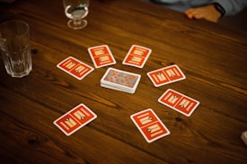 A wooden table with several playing cards arranged in a circle around a stack of cards in the center. Each card has a red back with text and illustrations. Two drinking glasses, one with a clear liquid, are also on the table, with a person's arm visible in the background.