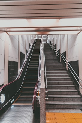 An indoor setting featuring an escalator on the left and a parallel staircase on the right, both leading upward. The walls are lined with light-colored tiles, and there's a ceiling with horizontal panels. The area appears clean and modern, with symmetrical architectural elements.