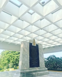 A large marble monument with engraved text stands under a geometric ceiling made of square skylights. Green bushes and trees surround the area, creating a contrasting natural backdrop.