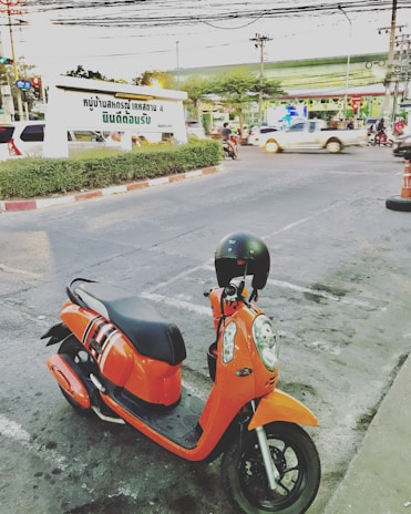 A bright orange scooter is parked on the side of a busy street. The scooter has a black seat and a helmet placed on its handlebar. In the background, there are vehicles passing by and a large billboard with Thai text. There’s a mix of cars and motorcycles on the road and various businesses visible in the background.