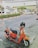 A bright orange scooter is parked on the side of a busy street. The scooter has a black seat and a helmet placed on its handlebar. In the background, there are vehicles passing by and a large billboard with Thai text. There’s a mix of cars and motorcycles on the road and various businesses visible in the background.