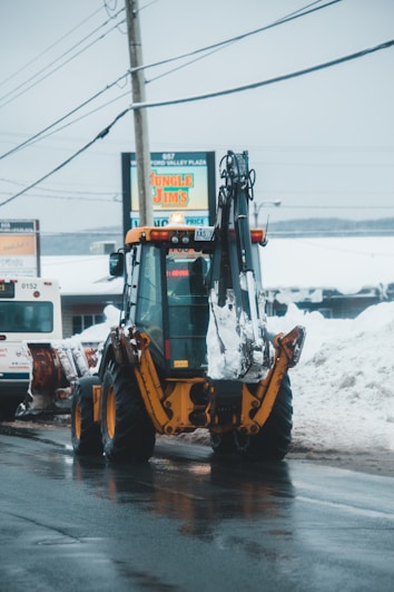 A yellow backhoe loader is parked on a wet street with snow piled on either side. Overhead power lines stretch across the scene, and in the background, a sign for a business called 'Jungle Jim's' is visible along with other buildings and vehicles.