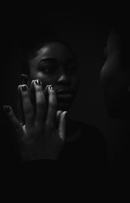 A person is gazing at their reflection in a mirror with their hand pressed against the glass, creating a powerful sense of introspection. The image is captured in low light, emphasizing shadows and creating a dramatic, contemplative atmosphere.