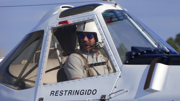 A man wearing a helmet and sunglasses is seated inside the cockpit of an aircraft. The word 'RESTRINGIDO' is visible on the exterior of the cockpit. The cockpit windows are large and transparent, allowing a clear view of the interior. The sky is a light blue in the background, and some vegetation can be seen.
