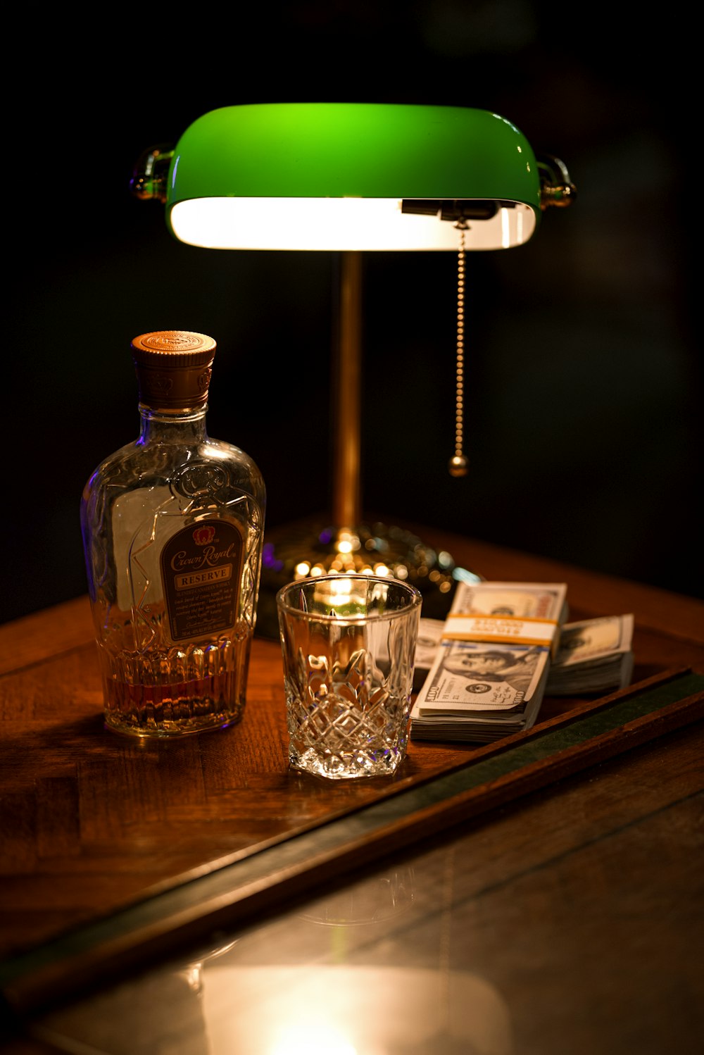 decanter by empty drinking glass cup by turned-on banker's lamp