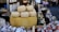 A rustic market stall displays an array of artisanal cheeses, including a large wheel of Parmesan cheese prominently placed with several smaller rounds stacked on top. Nearby are assorted salamis wrapped in various packaging. Bottles of olive oil, jars of preserved goods, and other gourmet items fill the background, creating a rich, gourmet setting.