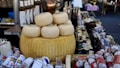 A rustic market stall displays an array of artisanal cheeses, including a large wheel of Parmesan cheese prominently placed with several smaller rounds stacked on top. Nearby are assorted salamis wrapped in various packaging. Bottles of olive oil, jars of preserved goods, and other gourmet items fill the background, creating a rich, gourmet setting.