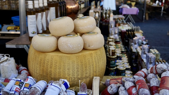 A rustic market stall displays an array of artisanal cheeses, including a large wheel of Parmesan cheese prominently placed with several smaller rounds stacked on top. Nearby are assorted salamis wrapped in various packaging. Bottles of olive oil, jars of preserved goods, and other gourmet items fill the background, creating a rich, gourmet setting.