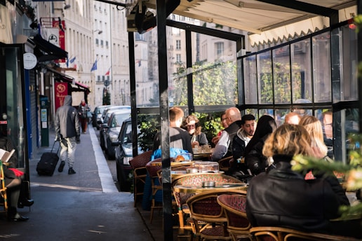 A bustling street scene in a European city with people dining at an outdoor café. The area is filled with parked cars along the sidewalk, and a pedestrian walks by pulling a suitcase. Sunlight filters through the café's transparent walls, illuminating the patrons enjoying their meals.