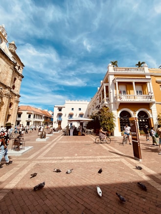 A lively town square with people walking and cycling. The area features colonial architecture, with colorful buildings, arched windows, and decorative balconies. Pigeons are scattered on the patterned cobblestone paving. The sky is clear with a few clouds, giving a warm and inviting atmosphere.