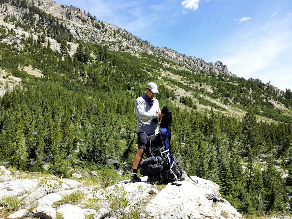 man wearing gray jacket with black and blue hiking backpack standing on rock viewing mountain under blue and white sky