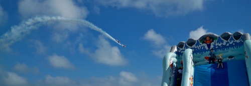 An inflatable slide resembling an underwater theme park sits under a bright blue sky. A plane performing an aerial maneuver leaves a white smoke trail curving across the sky.