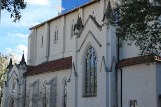 A large, white stone church with Gothic architectural features such as pointed arches and decorative elements. The building has a red-tiled roof and tall, narrow windows. Trees are partially visible, providing a natural frame on the sides.