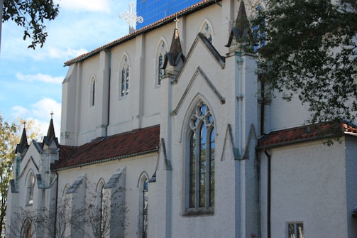 A large, white stone church with Gothic architectural features such as pointed arches and decorative elements. The building has a red-tiled roof and tall, narrow windows. Trees are partially visible, providing a natural frame on the sides.