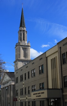 A tall church steeple rises prominently against a bright blue sky, adorned with a cross at its peak. The building's architecture features classic elements such as columns and brick facades, typical of traditional church designs. The signage indicates the name of the church, 'Downtown Baptist Church of Orlando', positioned on a rectangular section at the building's entrance. A street sign reads 'WRONG WAY' in red at the bottom left corner.