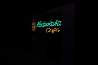 A neon sign with the words 'Karlsplatz Cafe' in bright green and yellow colors, mounted on a dark background.