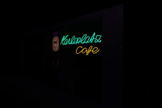 A neon sign with the words 'Karlsplatz Cafe' in bright green and yellow colors, mounted on a dark background.
