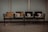 Four worn-out wooden chairs are aligned in a row, attached to a metal frame. They are situated in front of a plain, light-colored wall, likely in a public waiting area or a similar space. The floor is made of a gray material that could be concrete or tile.