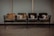 Four worn-out wooden chairs are aligned in a row, attached to a metal frame. They are situated in front of a plain, light-colored wall, likely in a public waiting area or a similar space. The floor is made of a gray material that could be concrete or tile.