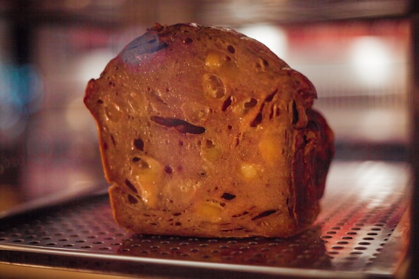 A loaf of bread with a dense texture, featuring visible chunks of nuts and possibly fruit. The surface has a rich, golden-brown color and is set against a blurred background with warm lighting.