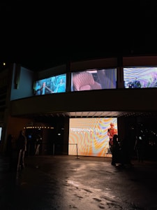 A nighttime scene outside a movie theater displaying advertisements on large digital screens. The marquee prominently displays 'TERMINATOR'. Shadows of people can be seen standing around the entrance, with the ground wet and reflecting light.