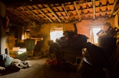 A dimly lit attic space appears cluttered with various items such as cardboard boxes, wooden crates, old furniture, and bundles of fabric. Dusty beams of sunlight filter through a small window, casting a warm glow across the exposed wooden rafters and stone walls. The scene conveys a sense of abandonment and nostalgia, with cobwebs visible in the corners and items haphazardly stacked around.