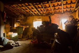 A dimly lit attic space appears cluttered with various items such as cardboard boxes, wooden crates, old furniture, and bundles of fabric. Dusty beams of sunlight filter through a small window, casting a warm glow across the exposed wooden rafters and stone walls. The scene conveys a sense of abandonment and nostalgia, with cobwebs visible in the corners and items haphazardly stacked around.