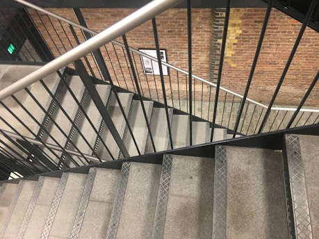 A staircase constructed with metal railings and stone steps, enclosed by a brick wall with visible pattern variations. A safety exit sign is seen on the left, and a wall-mounted notice is in the distance.