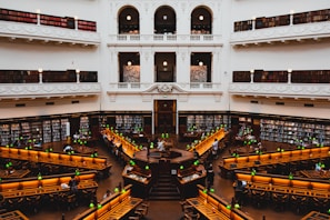 A grand library with high ceilings and ornate architecture, featuring rows of long wooden desks and numerous green desk lamps. The spacious room is surrounded by several tiers of balconies filled with bookshelves. People are scattered throughout the library, engaged in reading and studying.