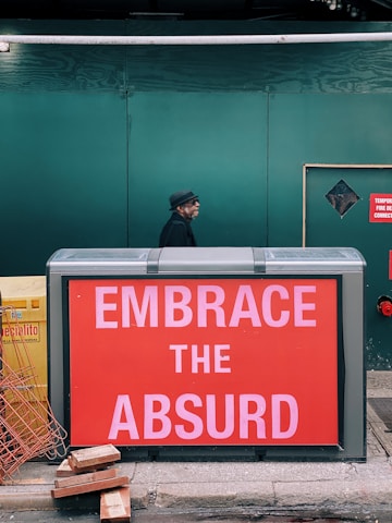 A man wearing a hat and dark clothing walks past a large red sign with the text 'EMBRACE THE ABSURD'. The background consists of a green construction barrier and there are construction materials like bricks and metal wiring on the ground.