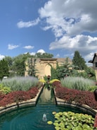 A lush garden with a symmetrical design features a long rectangular pond lined with red and green shrubbery. The water in the pond is clear, with a small fountain creating ripples at its end. Various plants surround the path leading towards an arched stone structure at the back. The sky above is blue with white, fluffy clouds, and trees and plants are abundant, adding to the serene and peaceful atmosphere.
