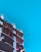 A partly constructed building with visible scaffolding in bright pink is set against a clear blue sky. The construction appears to be in its early stages with exposed concrete floors and some construction materials on site.