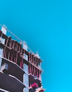 A partly constructed building with visible scaffolding in bright pink is set against a clear blue sky. The construction appears to be in its early stages with exposed concrete floors and some construction materials on site.