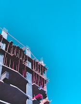 A partly constructed building with visible scaffolding in bright pink is set against a clear blue sky. The construction appears to be in its early stages with exposed concrete floors and some construction materials on site.