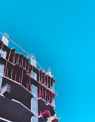 A partly constructed building with visible scaffolding in bright pink is set against a clear blue sky. The construction appears to be in its early stages with exposed concrete floors and some construction materials on site.