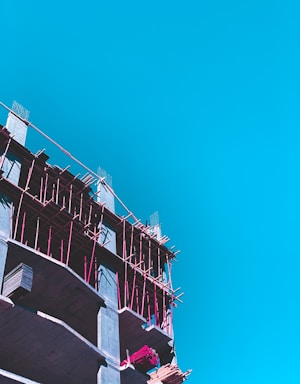 A partly constructed building with visible scaffolding in bright pink is set against a clear blue sky. The construction appears to be in its early stages with exposed concrete floors and some construction materials on site.