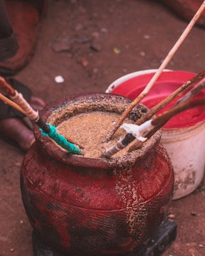 A large, red container filled with some kind of grain or powder with several sticks inserted into it. The container looks old and worn. In the background, there is another white and red container on a dirt surface.