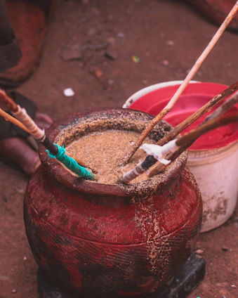 A large, red container filled with some kind of grain or powder with several sticks inserted into it. The container looks old and worn. In the background, there is another white and red container on a dirt surface.