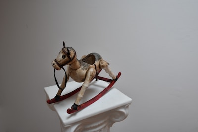 A vintage wooden rocking horse with a weathered finish sits atop a white ornate pedestal. The horse has a rustic appearance with worn paint and visible age marks. It features red rockers and black stirrups, standing out against the minimalist background.