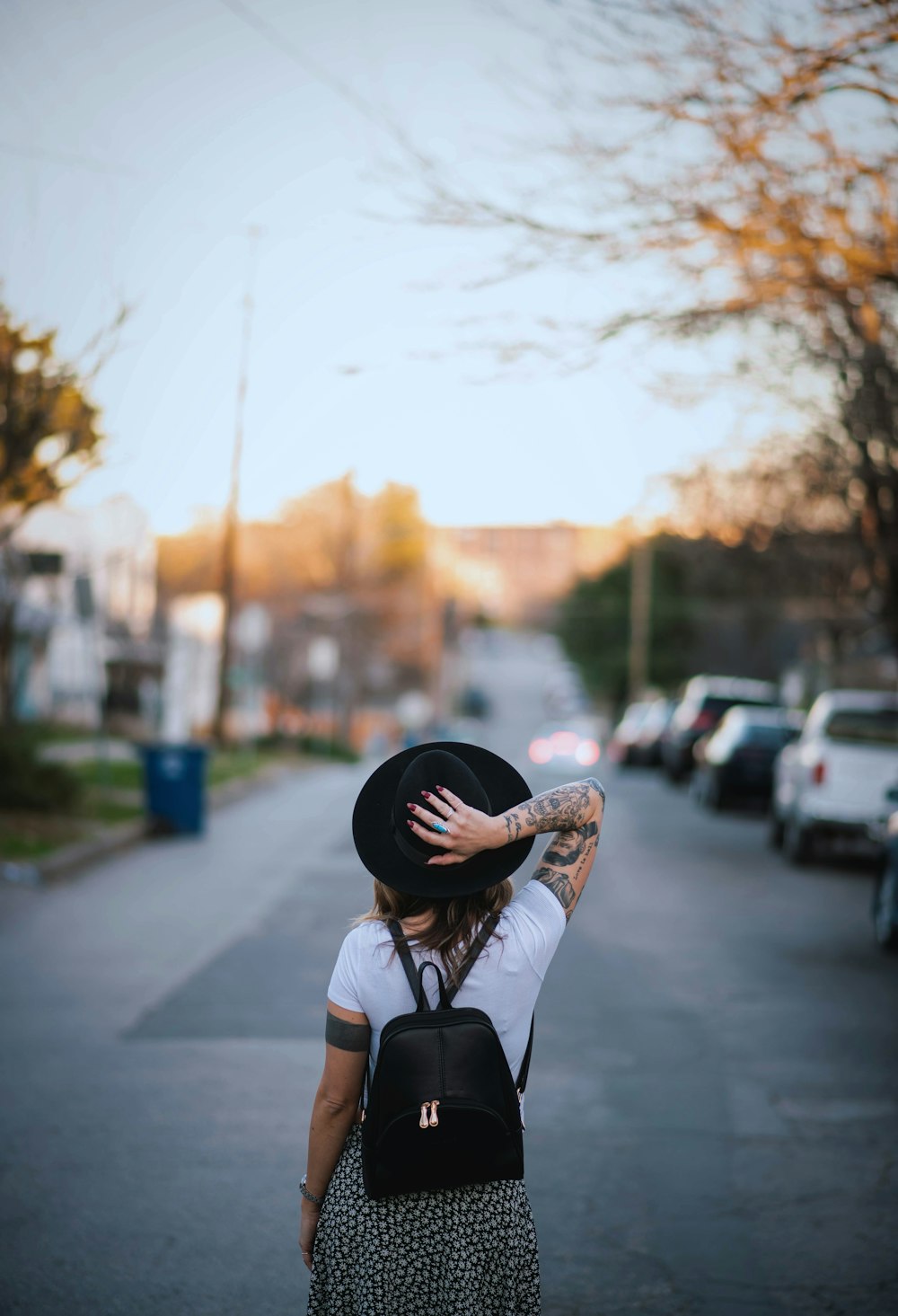 woman wearing white crew-neck t-shirt and black hat with black leather backpack standing while facing back on road during daytime