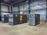 A spacious industrial facility houses several large air compressors. The machines are labeled with the brand 'Atlas Copco' and feature different series codes including 'Z' and 'G'. Blue and green piping connects the compressors, indicating an air or fluid handling system. The walls and ceiling are made of corrugated metal, adding to the utilitarian appearance. The floor is made of smooth concrete.