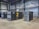 A spacious industrial facility houses several large air compressors. The machines are labeled with the brand 'Atlas Copco' and feature different series codes including 'Z' and 'G'. Blue and green piping connects the compressors, indicating an air or fluid handling system. The walls and ceiling are made of corrugated metal, adding to the utilitarian appearance. The floor is made of smooth concrete.