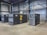A spacious industrial facility houses several large air compressors. The machines are labeled with the brand 'Atlas Copco' and feature different series codes including 'Z' and 'G'. Blue and green piping connects the compressors, indicating an air or fluid handling system. The walls and ceiling are made of corrugated metal, adding to the utilitarian appearance. The floor is made of smooth concrete.