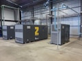 A spacious industrial facility houses several large air compressors. The machines are labeled with the brand 'Atlas Copco' and feature different series codes including 'Z' and 'G'. Blue and green piping connects the compressors, indicating an air or fluid handling system. The walls and ceiling are made of corrugated metal, adding to the utilitarian appearance. The floor is made of smooth concrete.