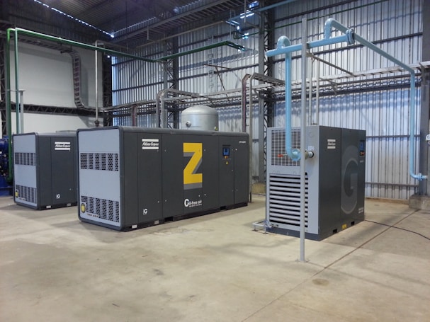 A spacious industrial facility houses several large air compressors. The machines are labeled with the brand 'Atlas Copco' and feature different series codes including 'Z' and 'G'. Blue and green piping connects the compressors, indicating an air or fluid handling system. The walls and ceiling are made of corrugated metal, adding to the utilitarian appearance. The floor is made of smooth concrete.