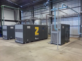 A spacious industrial facility houses several large air compressors. The machines are labeled with the brand 'Atlas Copco' and feature different series codes including 'Z' and 'G'. Blue and green piping connects the compressors, indicating an air or fluid handling system. The walls and ceiling are made of corrugated metal, adding to the utilitarian appearance. The floor is made of smooth concrete.