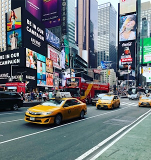 A bustling urban street scene with several iconic yellow taxis driving down a busy avenue. Bright, colorful advertisements and large digital billboards cover the facades of tall buildings. A double-decker tour bus and numerous pedestrians are visible, conveying a sense of vibrant city life.